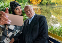 Wilsberg (Leonard Lansink) sitzt neben Bonnie von Laer (Johanna Ingelfinger) auf einer bunten Parkbank vor einem kleinen See. Bonnie holt ihr Handy vor sich und Wilsberg, um ein Selfie zu machen. Beide lächeln in die Handy-Kamera.