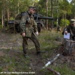 Soldaten sitzen bei einer Übung im Wald, im Hintergrund ein Leopard2-Panzer