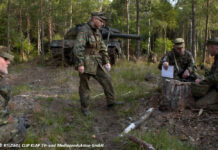 Soldaten sitzen bei einer Übung im Wald, im Hintergrund ein Leopard2-Panzer