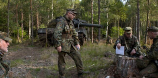 Soldaten sitzen bei einer Übung im Wald, im Hintergrund ein Leopard2-Panzer