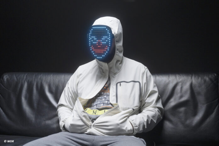 mensch in Kapuzenpulli und mit Maske sitzt auf einem Sofa vor schwarzem Hintergrund