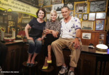 Susanne Daubner, Ina Müller und Götz Otto sitzen auf einem Tisch in einer Kneipe und schauen in die Kamera