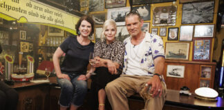 Susanne Daubner, Ina Müller und Götz Otto sitzen auf einem Tisch in einer Kneipe und schauen in die Kamera