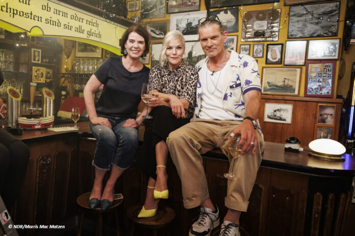 Susanne Daubner, Ina Müller und Götz Otto sitzen auf einem Tisch in einer Kneipe und schauen in die Kamera