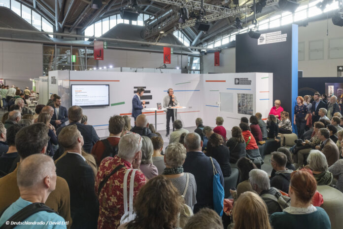 Ein Stand des Deutschlandradios auf der Frankfurter Buchmesse, Publikum hört einem stehenden Redner zu