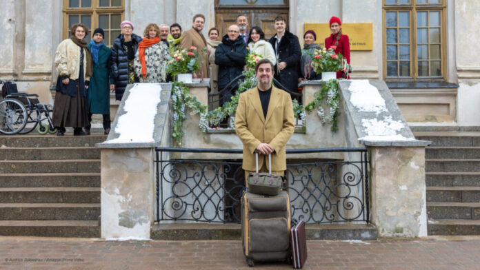 Sebastian Pastewka mit Koffer vor Schlosstreppe, auf der Hochzeitsgesellschaft steht. Aus Prime Video-Film "Die Brautentführung" (AT)